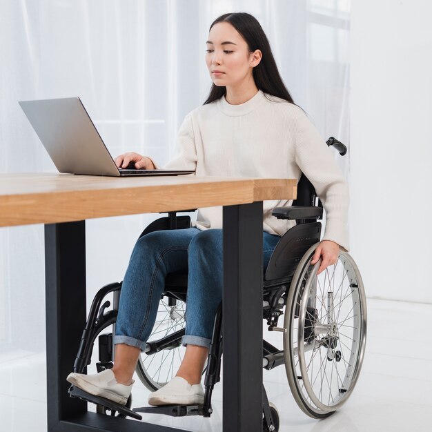 Giovane donna invalida che si siede sulla sedia a rotelle facendo uso del computer portatile