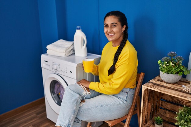 Giovane donna ispanica che beve caffè in attesa della lavatrice in lavanderia