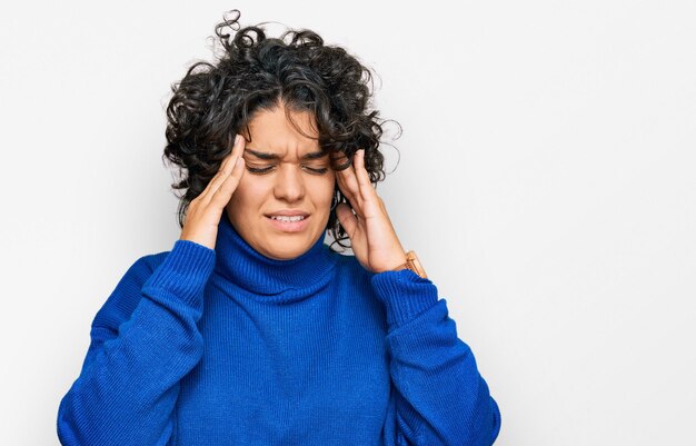 Giovane donna ispanica con i capelli ricci che indossa un maglione a collo alto con la mano sulla testa mal di testa perché lo stress soffre di emicrania