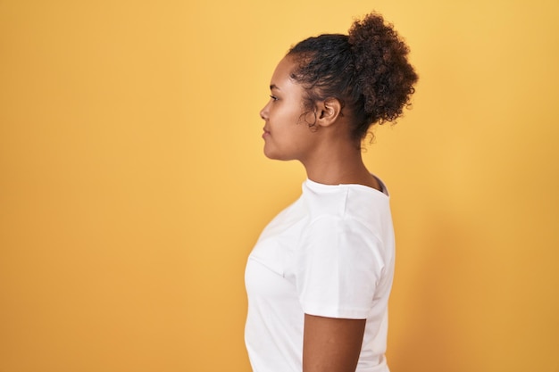 Giovane donna ispanica con i capelli ricci in piedi su sfondo giallo che guarda al lato, rilassa la posa del profilo con il viso naturale e il sorriso fiducioso.