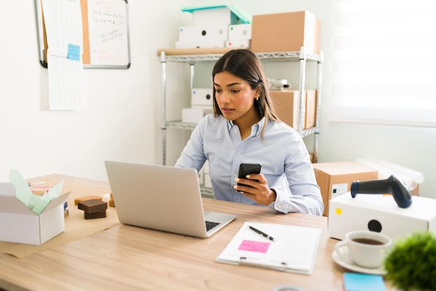 Giovane donna ispanica nel suo ufficio che guarda il suo laptop e smartphone per controllare gli ordini dei clienti sul suo negozio di bellezza online