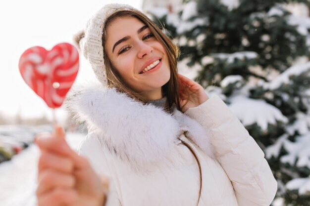 Giovane donna ispirata in cappello lavorato a maglia bianco che si diverte con il lecca-lecca cuore rosa sulla strada piena di neve. Donna attraente con la caramella in posa con il sorriso nella mattina congelata.