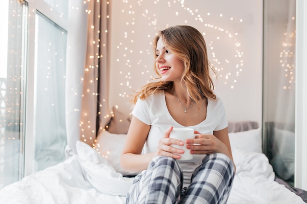 Giovane donna ispirata in maglietta bianca che guarda meraviglia e beve caffè. Tiro al coperto del bellissimo modello femminile in pantaloni a scacchi che si siede sul letto.