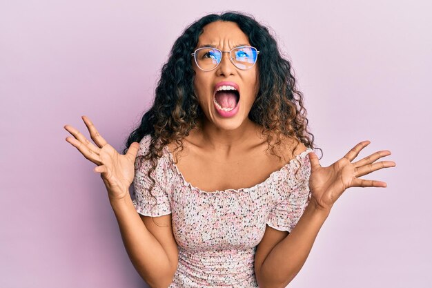 Giovane donna latina che indossa abiti casual e occhiali pazzi e pazzi che gridano e urlano con espressione aggressiva e braccia alzate. concetto di frustrazione.