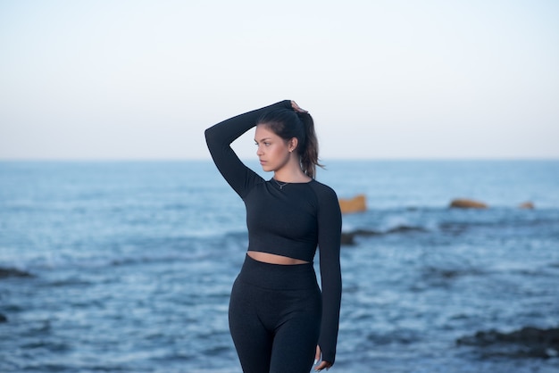 Giovane donna latina che indossa abiti da allenamento neri, in piedi sulla spiaggia e in posa