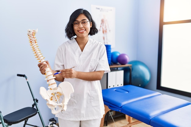 Giovane donna latina che indossa l'uniforme del fisioterapista che tiene il modello anatomico della colonna vertebrale presso la clinica di fisioterapia