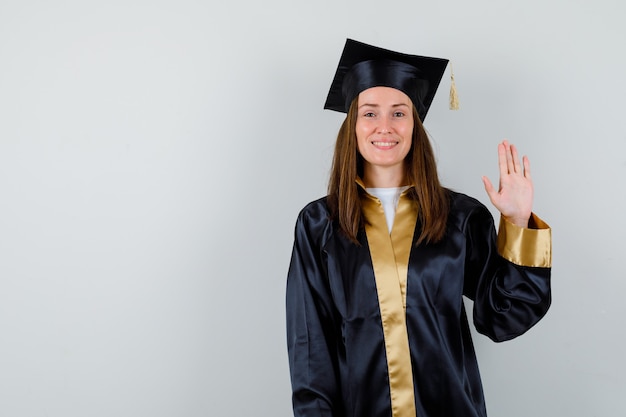 Giovane donna laureata che mostra la palma per il saluto in abito accademico e guardando allegro, vista frontale.
