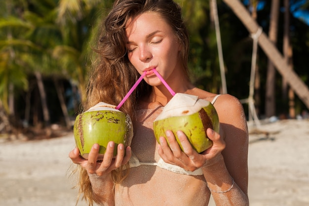 Giovane donna magra in costume da bagno bikini bianco con noci di cocco, sorridente, prendere il sole sulla spiaggia tropicale.