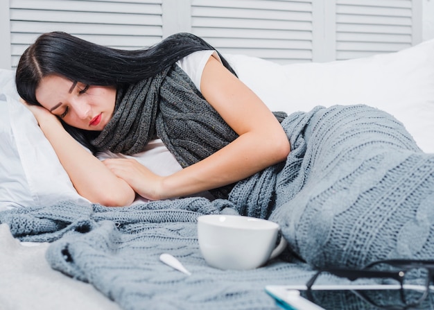 Giovane donna malata che dorme sul letto con il termometro; tazza; occhiali da vista e tavoletta digitale