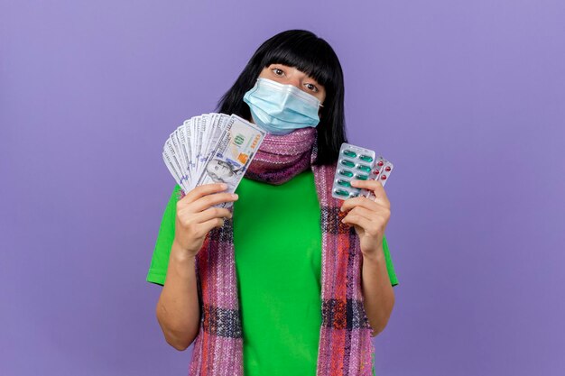 Giovane donna malata che indossa maschera e sciarpa tenendo i soldi e confezioni di capsule guardando la parte anteriore isolata sulla parete viola con lo spazio della copia