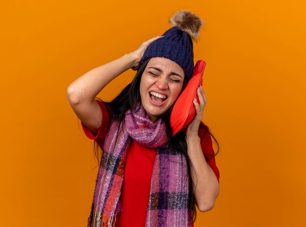 Giovane donna malata dolorante che indossa cappello invernale e sciarpa toccando il viso con la borsa dell'acqua calda tenendo la mano sulla testa con gli occhi chiusi isolati sulla parete arancione