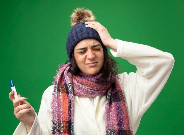 Giovane donna malata dolorante che indossa il cappello invernale della veste e la sciarpa che tiene il termometro che tiene la mano sulla testa con gli occhi chiusi isolati sulla parete verde