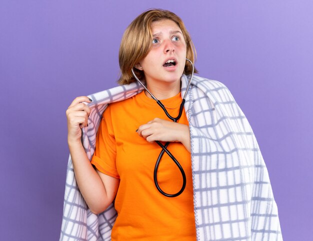 Giovane donna malsana avvolta in una coperta calda che si sente male ascoltando il suo battito cardiaco usando lo stetoscopio che sembra preoccupata in piedi sul muro viola