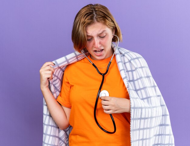 Giovane donna malsana avvolta in una coperta calda che si sente male ascoltando il suo battito cardiaco usando lo stetoscopio che sembra preoccupata in piedi sul muro viola