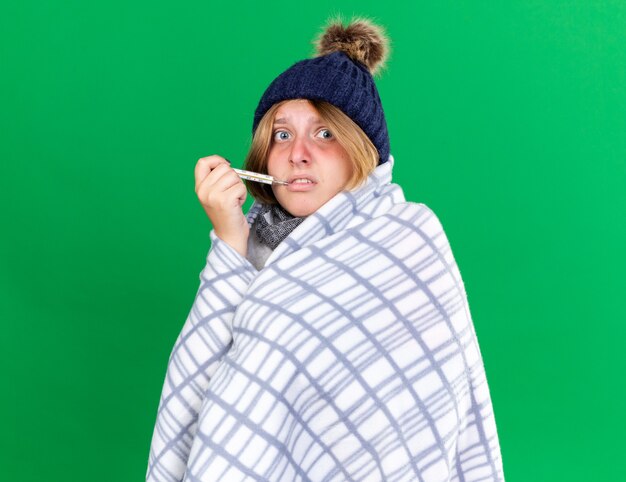 Giovane donna malsana avvolta in una coperta che indossa un cappello che misura la sua temperatura corporea usando un termometro che soffre di influenza con febbre che sembra preoccupata in piedi sul muro verde green