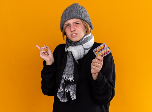 Giovane donna malsana che indossa un cappello caldo e con una sciarpa intorno al collo si sente male soffre di raffreddore e influenza tenendo pillole puntate con il dito indice sul lato in piedi sul muro arancione