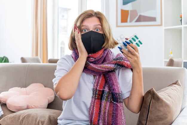 Giovane donna malsana con sciarpa calda intorno al collo con maschera protettiva per il viso sensazione di malessere e malati che soffrono di influenza e raffreddore che tengono pillole che sembrano confusi seduti sul divano nel soggiorno luminoso