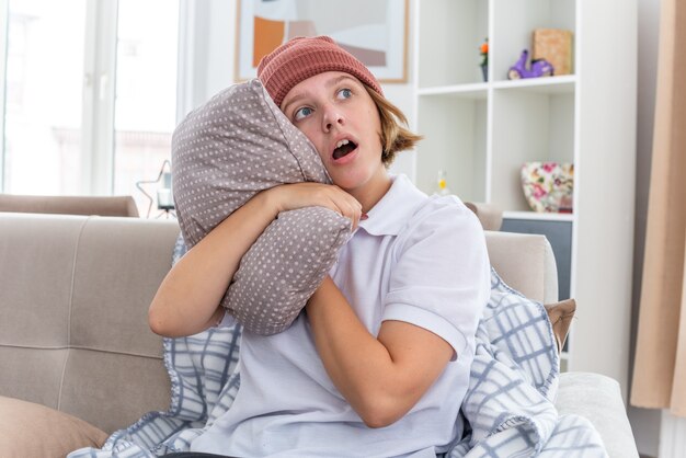 Giovane donna malsana in cappello caldo con coperta che sembra malata e malata che soffre di raffreddore e influenza che tiene cuscino che guarda da parte preoccupata seduta sul divano in un soggiorno luminoso