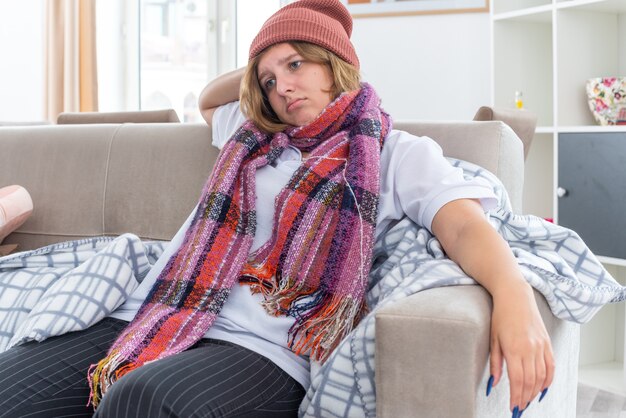 Giovane donna malsana in cappello caldo con sciarpa intorno al collo sensazione di malessere e malata che soffre di raffreddore e influenza che sembra preoccupata seduta sul divano in un soggiorno luminoso