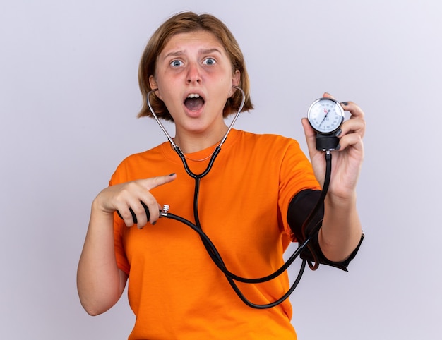Giovane donna malsana in maglietta arancione che si sente male misurando la pressione sanguigna usando il tonometro che sembra preoccupata in piedi sul muro bianco
