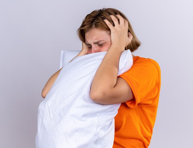 Giovane donna malsana in t-shirt arancione che tiene il cuscino che si sente male soffre di influenza con febbre e forte mal di testa in piedi sul muro bianco white