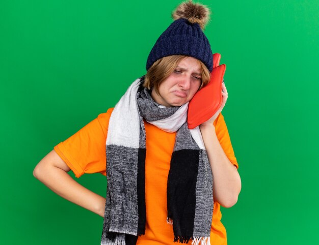 Giovane donna malsana in t-shirt arancione con cappello e sciarpa calda intorno al collo sensazione terribile con in mano una borsa dell'acqua calda che soffre di espressione fredda e triste