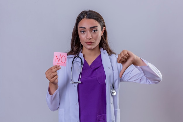 Giovane donna medico in camice bianco con fonendoscopio tenendo la carta promemoria senza messaggio che mostra antipatia con i pollici verso il basso su sfondo bianco isolato