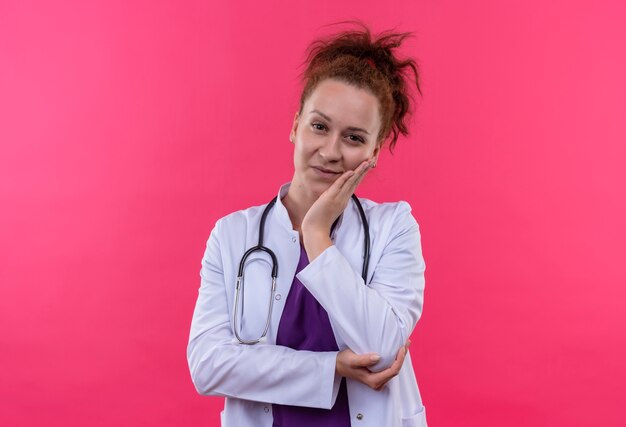 Giovane donna medico indossa camice bianco con lo stetoscopio con la mano sul mento guardando fiducioso in piedi oltre il muro rosa