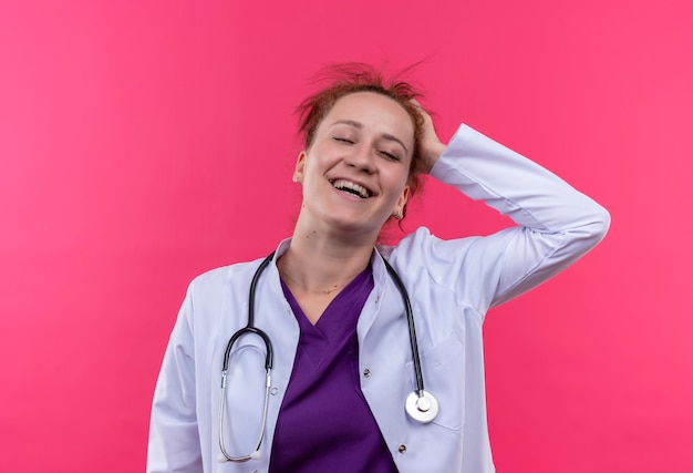Giovane donna medico indossa camice bianco con lo stetoscopio sorridente con la mano sulla testa felice e contento in piedi oltre il muro rosa