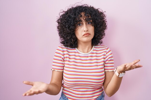 Giovane donna mediorientale in piedi su sfondo rosa all'oscuro e confusa con le braccia aperte, nessun concetto di idea.