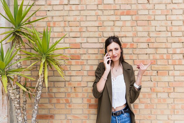 Giovane donna moderna che sta contro il muro di mattoni che parla sul dito indicante del cellulare