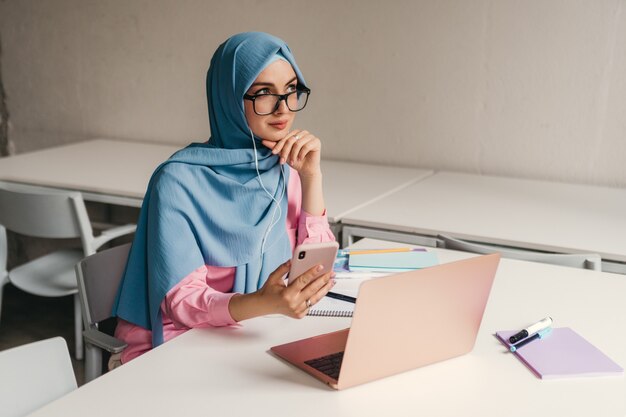 Giovane donna musulmana abbastanza moderna in hijab che lavora al computer portatile nella stanza dell'ufficio, istruzione online