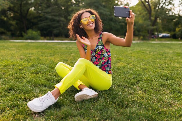 Giovane donna nera positiva alla moda che prende la foto del selfie che ascolta la musica sugli auricolari senza fili divertendosi nel parco, stile di modo di estate, vestito variopinto dei pantaloni a vita bassa