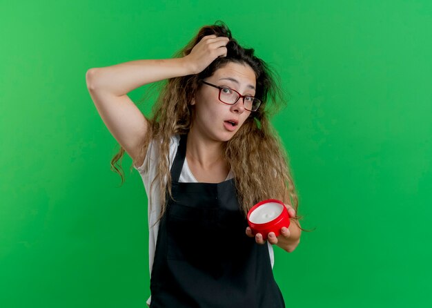 Giovane donna parrucchiere professionista in grembiule che tiene un barattolo di crema per capelli confusa con la mano sulla sua testa per errore