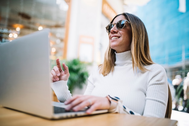 Giovane donna positiva pensierosa che utilizza computer portatile alla tavola in caffè della via