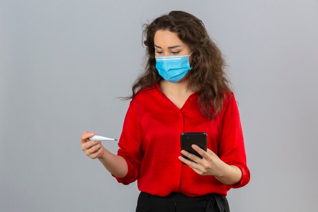 Giovane donna preoccupata che indossa camicetta rossa in maschera protettiva medica guardando il termometro digitale in mano mentre si tiene il cellulare in altra mano su sfondo bianco isolato