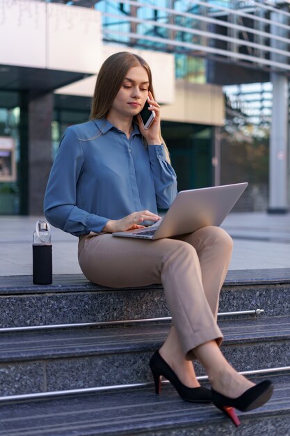 Giovane donna professionale che si siede sulla scala davanti all'edificio di vetro, tenendo il laptop in grembo e parlando al telefono cellulare