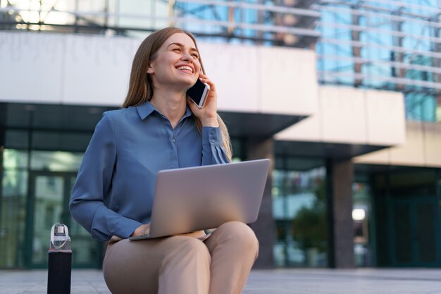 Giovane donna professionale che si siede sulla scala davanti all'edificio di vetro, tenendo il laptop in grembo e parlando al telefono cellulare