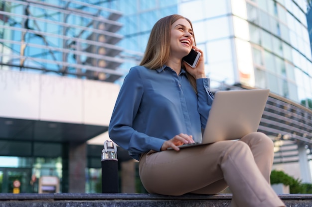 Giovane donna professionale che si siede sulla scala davanti all'edificio di vetro, tenendo il laptop in grembo e parlando al telefono cellulare
