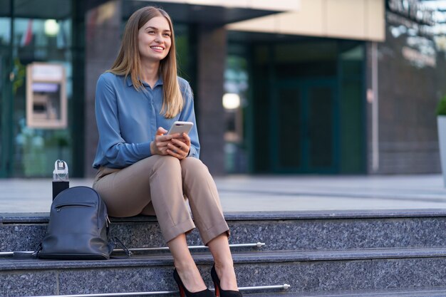 Giovane donna professionale seduta sulle scale di fronte al palazzo di vetro, parlando al telefono cellulare