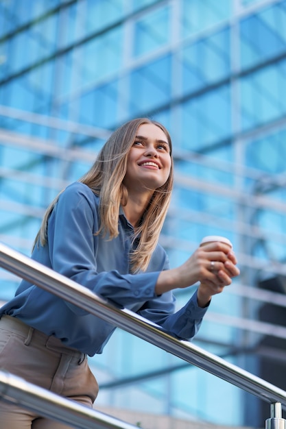 Giovane donna professionale sorridente con una pausa caffè durante la sua intera giornata lavorativa. Tiene un bicchiere di carta all'aperto vicino all'edificio aziendale mentre si rilassa e si gode la sua bevanda.