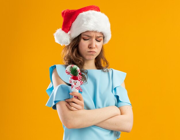 Giovane donna risentita in cima blu e cappello della Santa che tiene il bastoncino di zucchero di Natale che aggrotta le sopracciglia con le braccia incrociate
