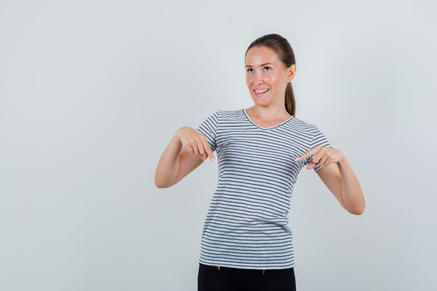 Giovane donna rivolta verso il basso e mostrando la lingua in maglietta, pantaloni e guardando speranzoso. vista frontale.