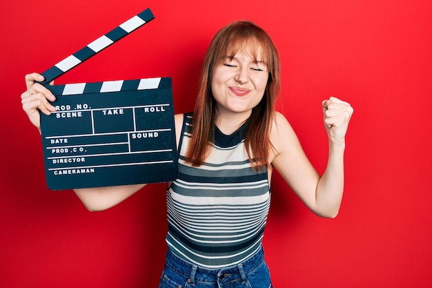 Giovane donna rossa che tiene l'assicella di un film video urlando orgogliosa, celebrando la vittoria e il successo molto eccitati con il braccio alzato