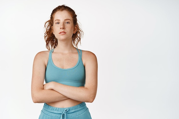 Giovane donna rossa sicura di sé con le braccia incrociate sul petto che sembra motivata Pareggiatore femminile che fa allenamento sportivo in palestra indossando uno sfondo bianco activewear