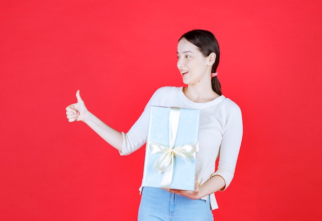 Giovane donna scioccata che tiene in mano una confezione regalo e fa un gesto con il pollice in su