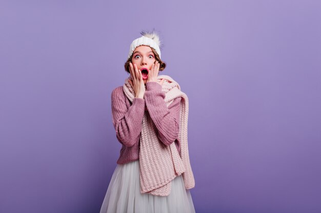 Giovane donna scioccata in cappello alla moda che grida sulla parete viola. Foto dell'interno della ragazza alla moda in sciarpa e maglione che tocca il fronte ed esprime stupore.