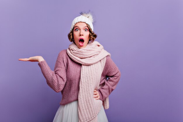 Giovane donna scioccata in sciarpa lavorata a maglia lunga che sta sulla parete viola con la bocca aperta. Ragazza in accessori invernali alla moda che esprimono emozioni sorprese.