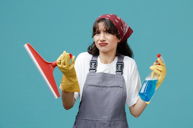 Giovane donna scontenta addetta alle pulizie che indossa bandana uniforme e guanti di gomma che mostrano tergicristallo e detergente guardando la fotocamera isolata su sfondo blu