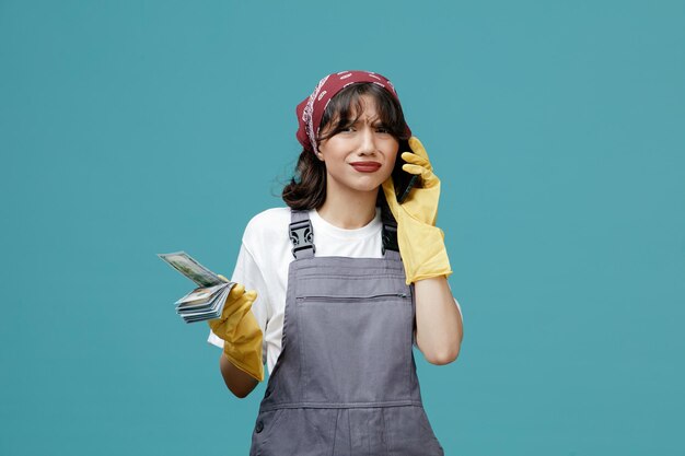 Giovane donna scontenta addetta alle pulizie che indossa bandana uniforme e guanti di gomma che tengono soldi guardando la fotocamera mentre si parla al telefono isolato su sfondo blu